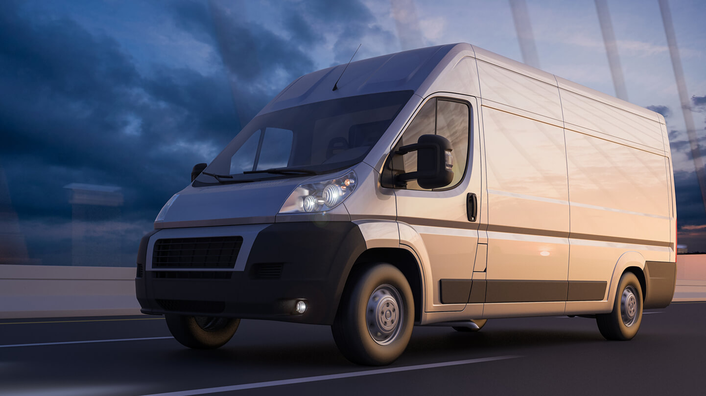 A white delivery van driving on a road at dusk with a cloudy sky in the background.