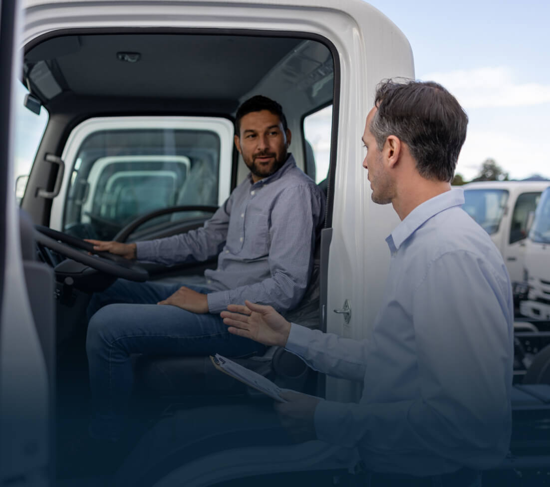 Two men discussing inside a truck; one sitting in the driver's seat, the other standing and gesturing with a clipboard.