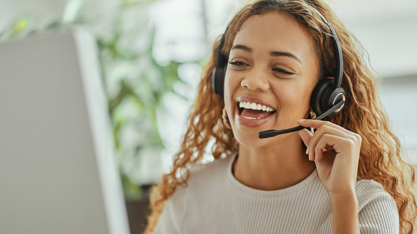 Woman smiling in a headset while on a call, sitting at a desk in front of a computer.