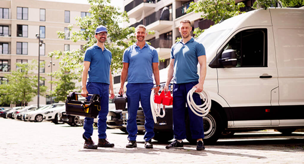 Three workers in blue uniforms stand by a white van, holding tools and hoses, with buildings in the background.