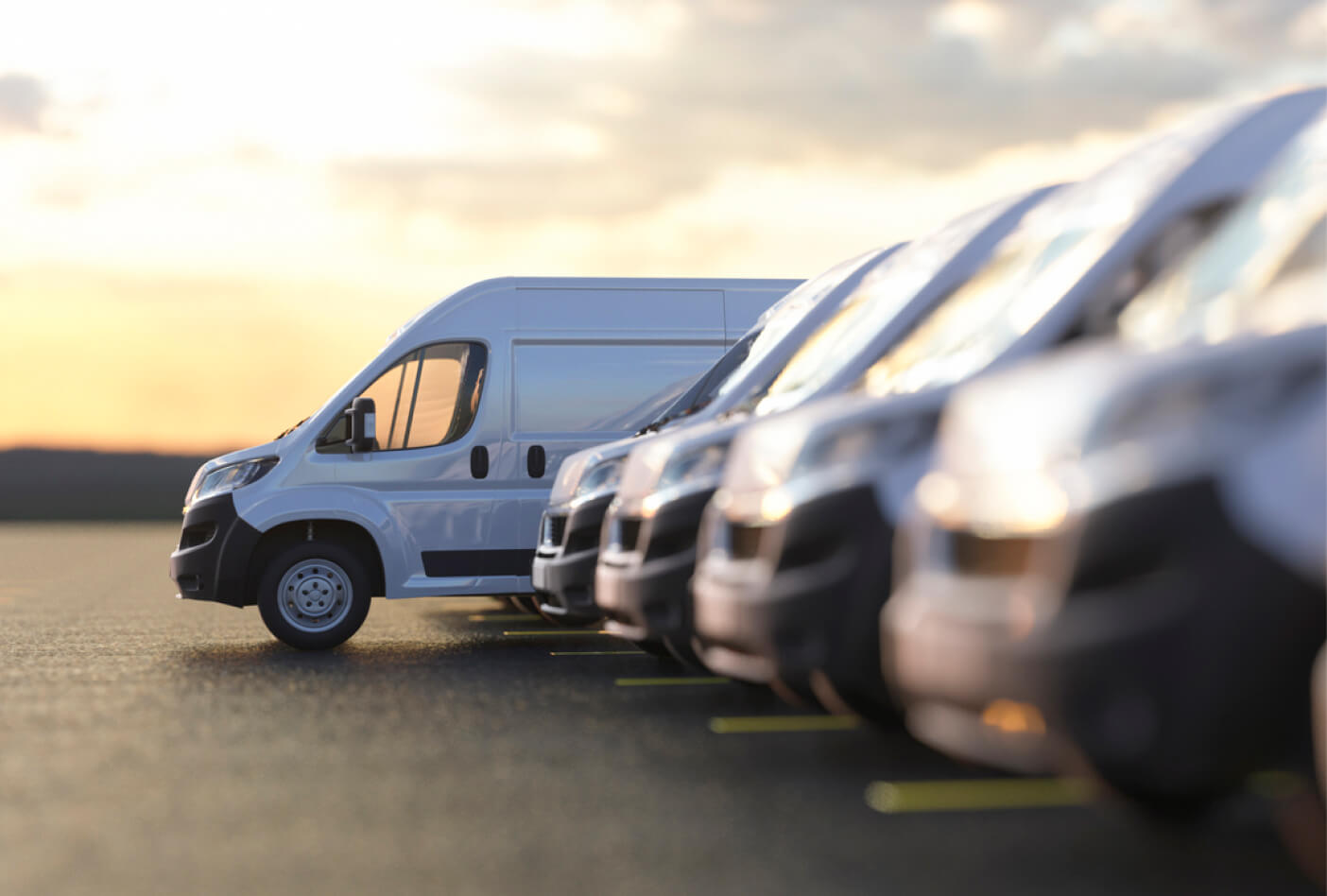 Row of white vans parked on a paved lot at sunset, with a clear sky in the background.