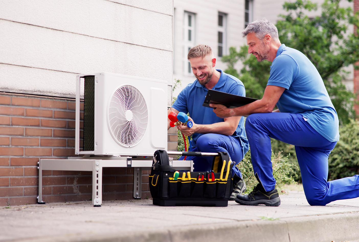 Two technicians in blue uniforms work on an outdoor air conditioning unit, with tools and a clipboard nearby.