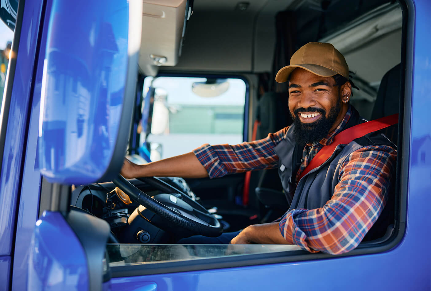 Driver smiling in a truck cab, wearing a brown cap and plaid shirt, with a safety belt fastened.