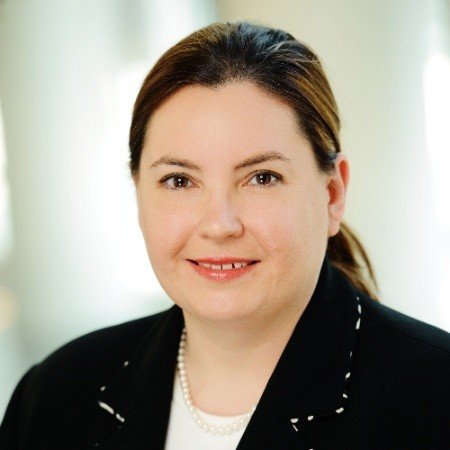 Woman with dark hair in a ponytail, wearing a black blazer and white shirt, smiling at the camera.