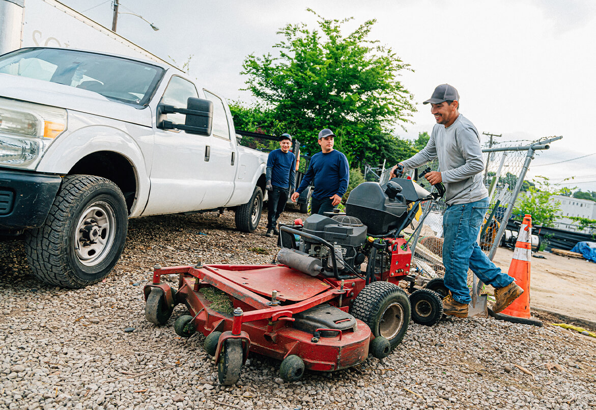 Three people work near a white truck; one operates a red lawn mower on a gravel surface.