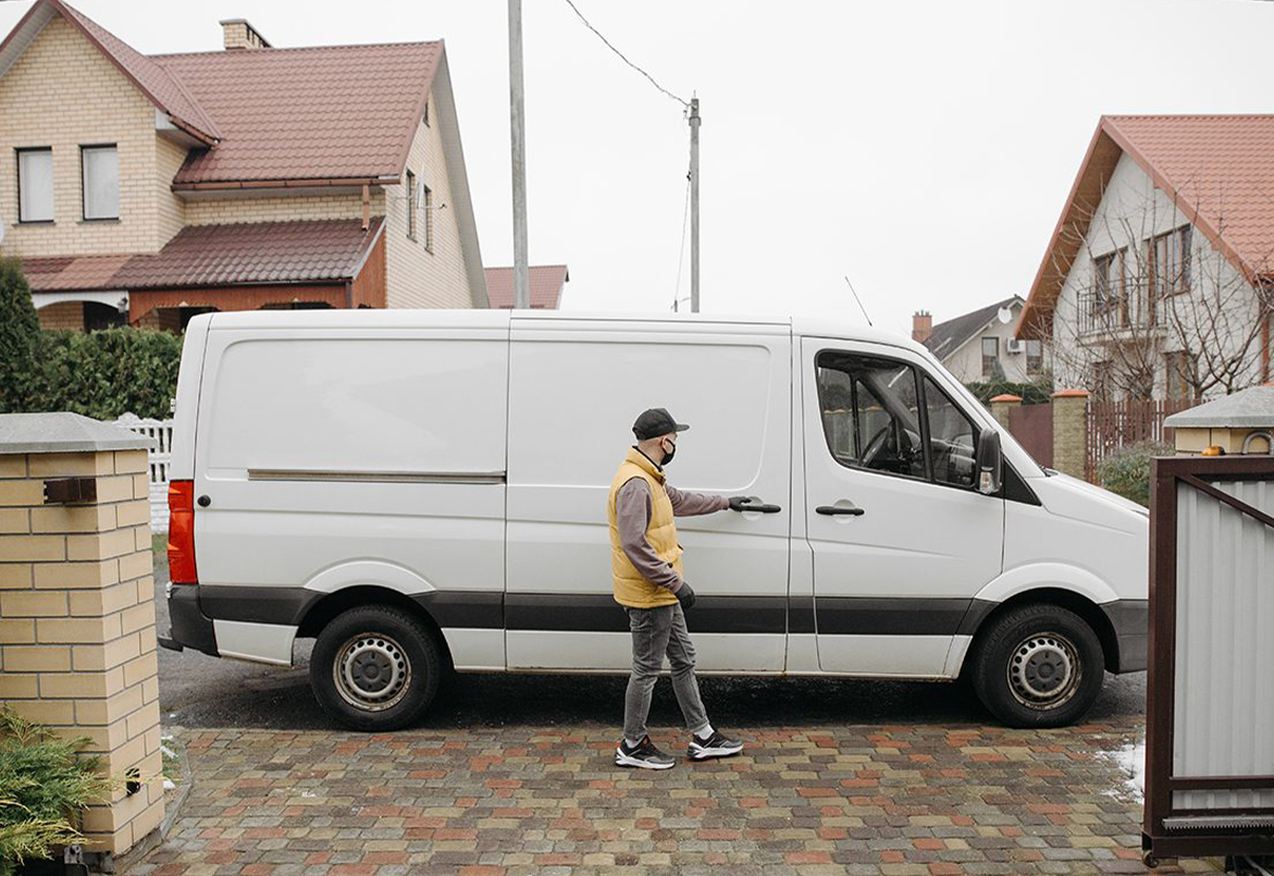 A man walks beside a white van parked in a residential neighborhood with brick houses.