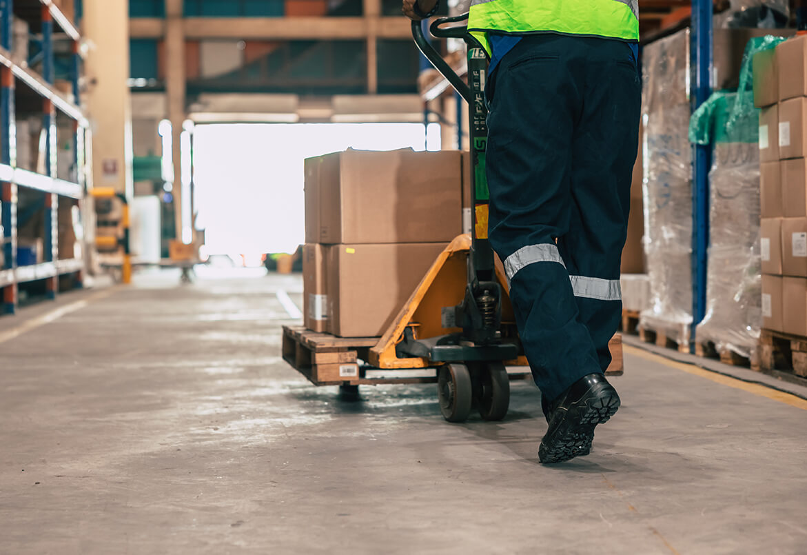 Person in safety gear using a hand pallet truck to move boxes in a warehouse aisle.