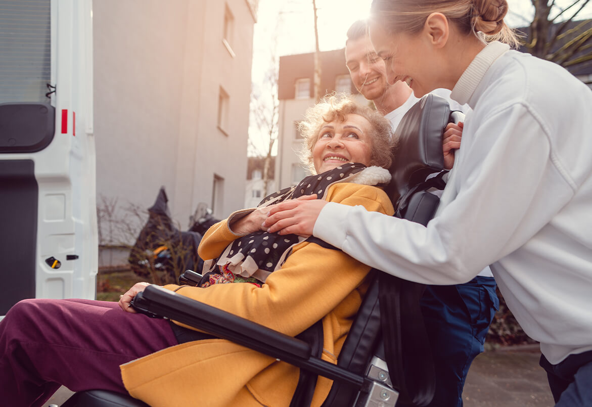 A smiling elderly woman in a wheelchair is assisted by two caregivers outdoors near a vehicle.