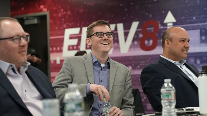 Three men in suits sit at a conference table, smiling and listening, with "ELEV8" in the background.