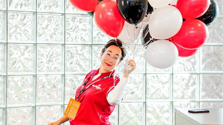 Person smiling, wearing a red shirt and name tag, holding a bunch of black, red, and white balloons indoors.