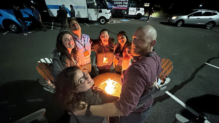 A group of people standing around a lit fire pit in a parking lot at night, smiling up at the camera.