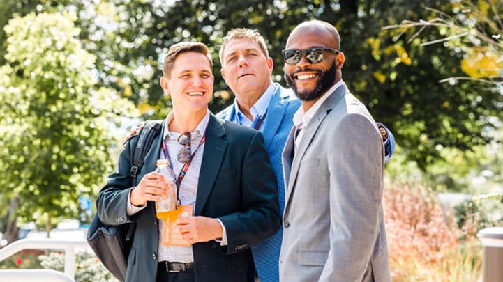 Three men in suits smiling outdoors; one holding a drink, another wearing sunglasses. Trees in the background.