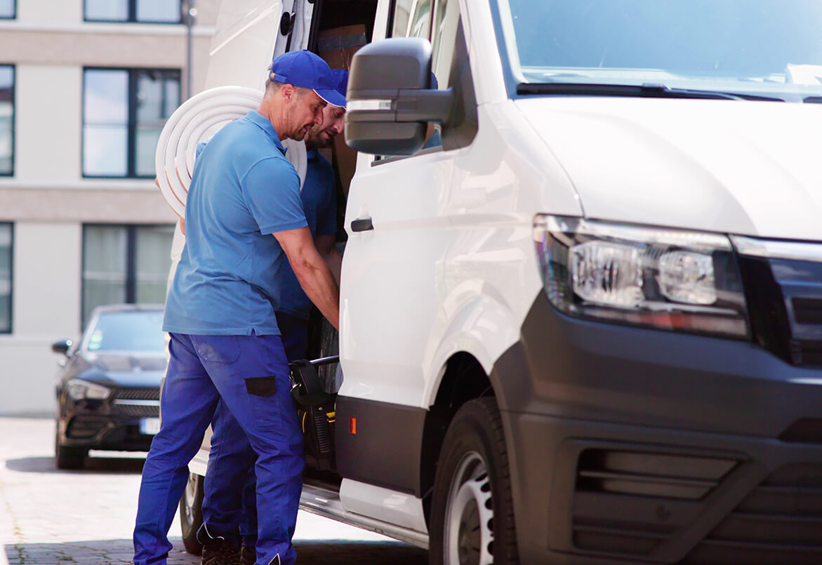 Two workers in blue uniforms load equipment into a white van parked on a sunny street.