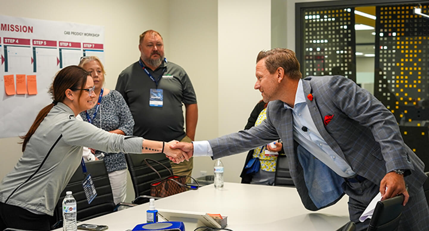 A group of people in a meeting room, with two individuals shaking hands across a table.