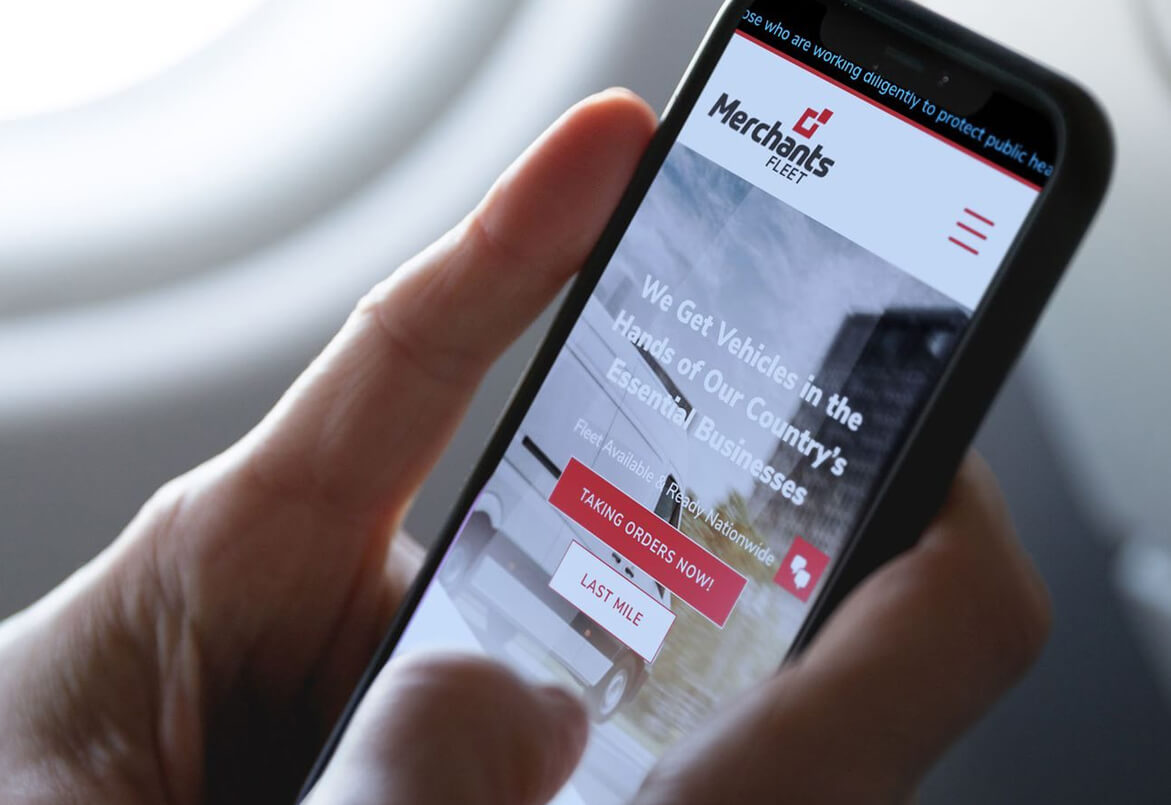 A person holds a smartphone displaying the Merchants Fleet website with buttons for taking orders and last mile services.