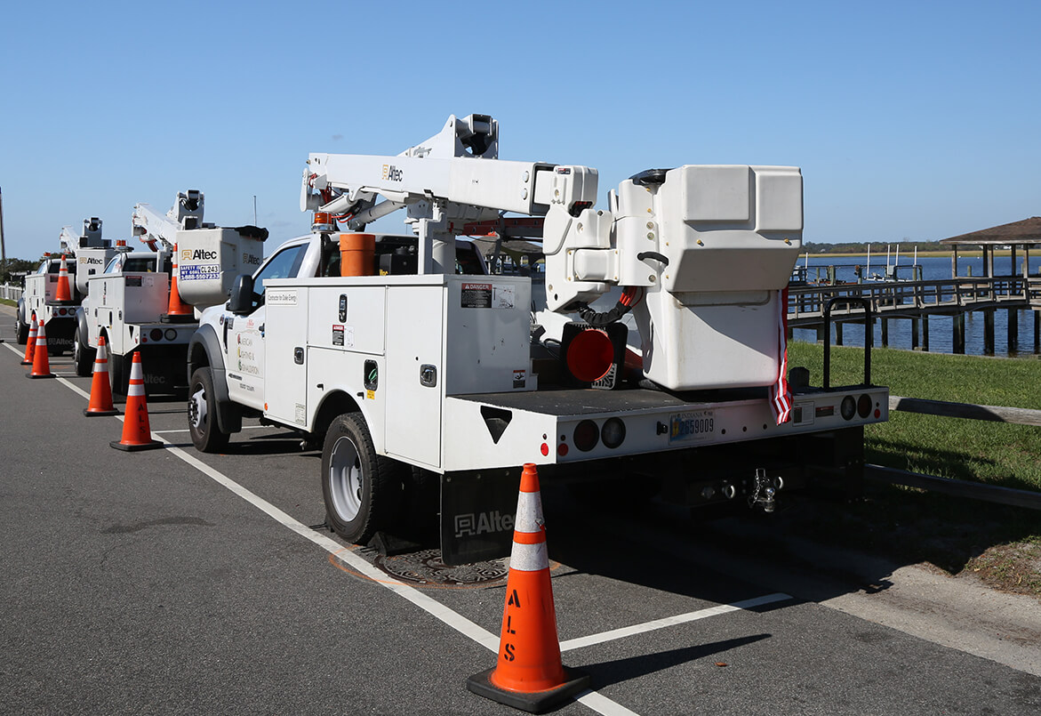 Utility trucks with crane arms parked by a road near water, surrounded by orange traffic cones.