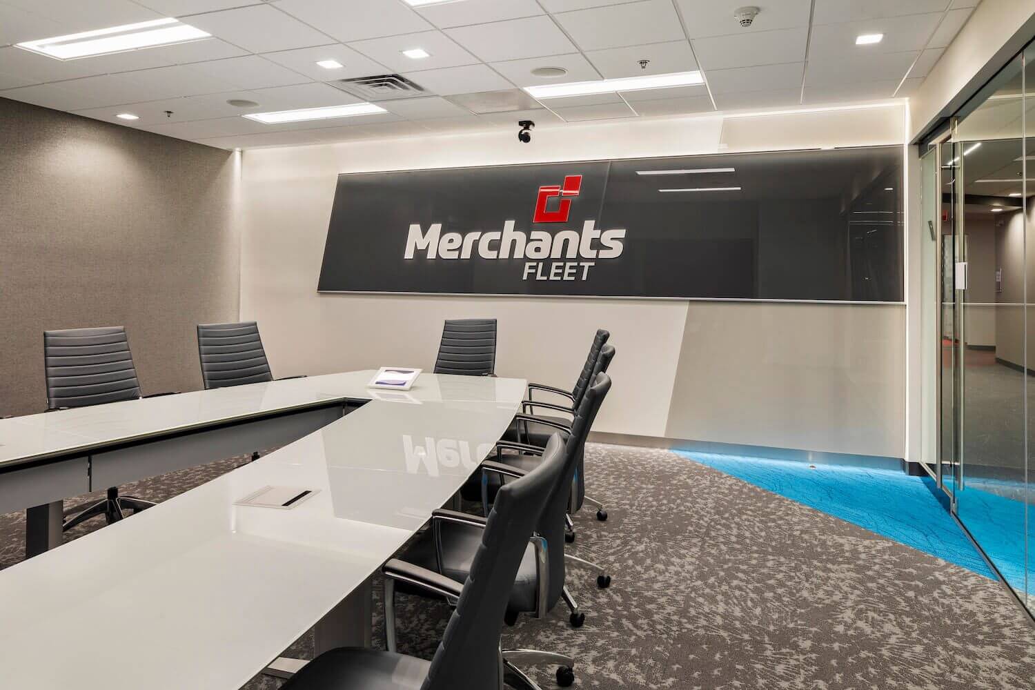 Modern conference room with a long glass table, black chairs, and a sign reading "Merchants Fleet" on the wall.
