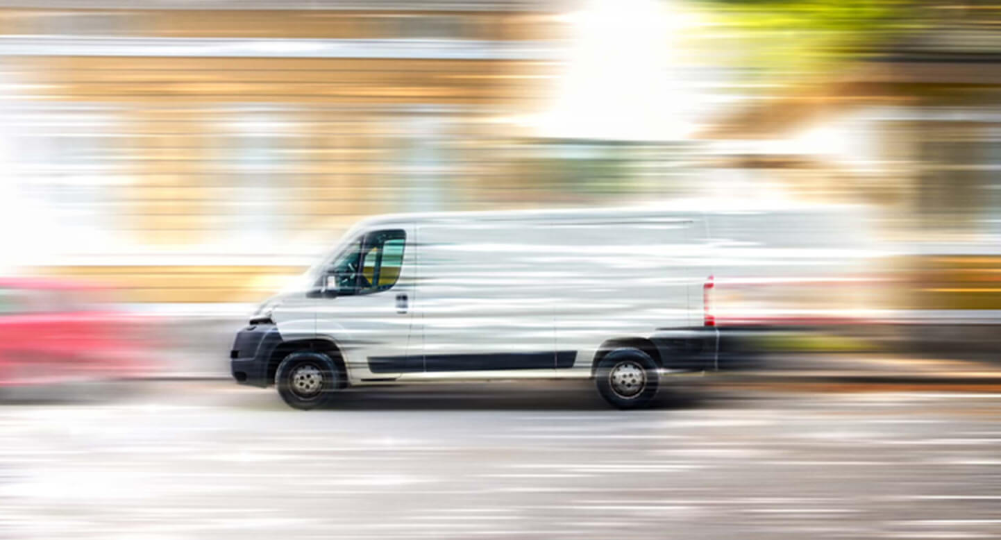 A white van speeds past on a blurred city street, with motion lines emphasizing its rapid movement.