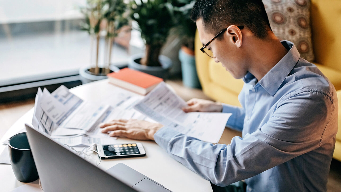 Man in blue shirt reviewing documents at a desk with a laptop, smartphone, and coffee mug nearby.