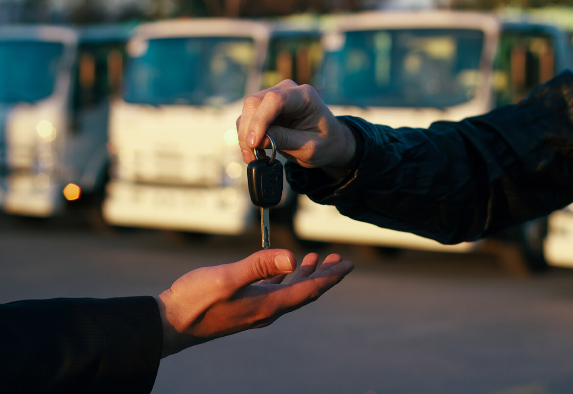 A hand gives a car key to another hand, with blurred vehicles in the background.