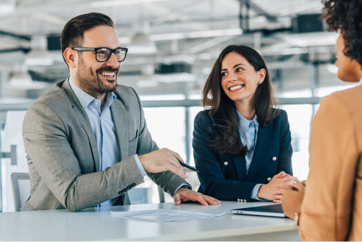 Two professionals in business attire smiling and conversing with a third person across a table in an office setting.