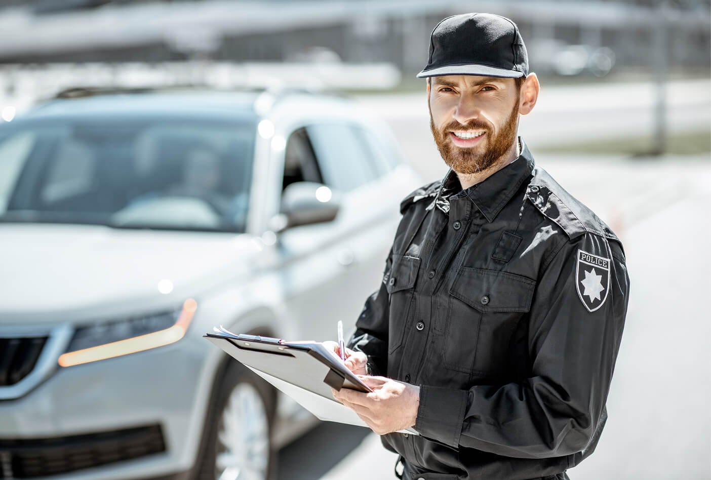 Smiling police officer with a clipboard stands near a gray vehicle on a sunny day.