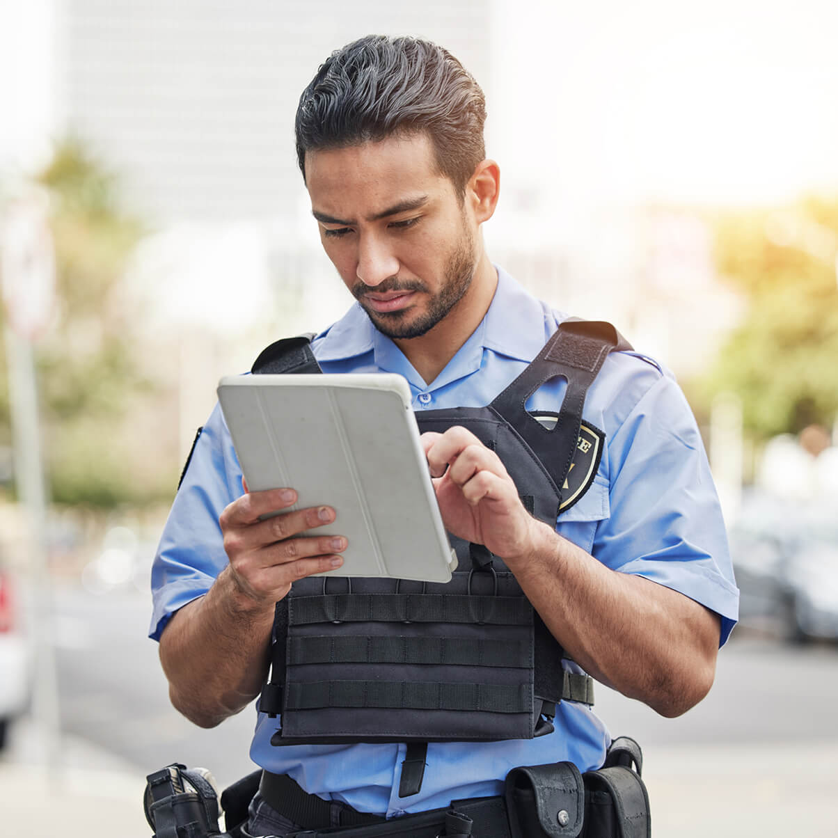 Police officer in uniform using a tablet outdoors, with buildings blurred in the background.