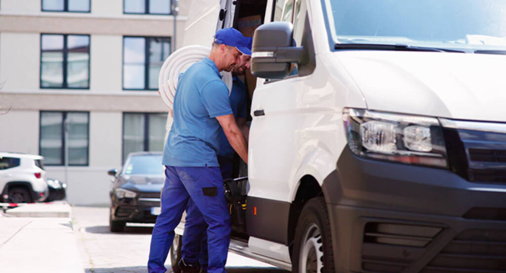 Delivery worker in blue uniform loading a white van on a sunny street with buildings in the background.