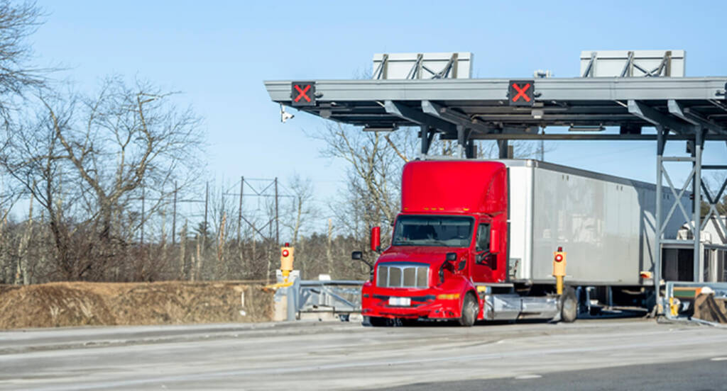 Red semi-truck passing through a toll booth on a clear day, with trees and bare land in the background.