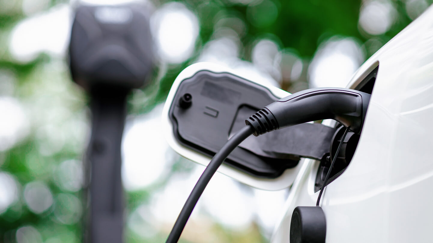 Close-up of an electric vehicle being charged at a charging station, with blurred greenery in the background.