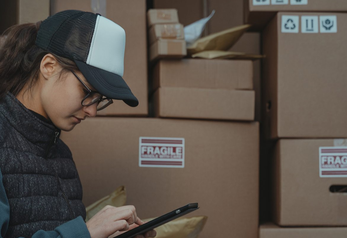 Person in a cap using a tablet, standing next to stacked boxes labeled "Fragile.