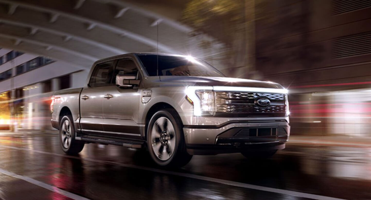 Silver pickup truck driving on a wet urban street at night with blurred city lights in the background.