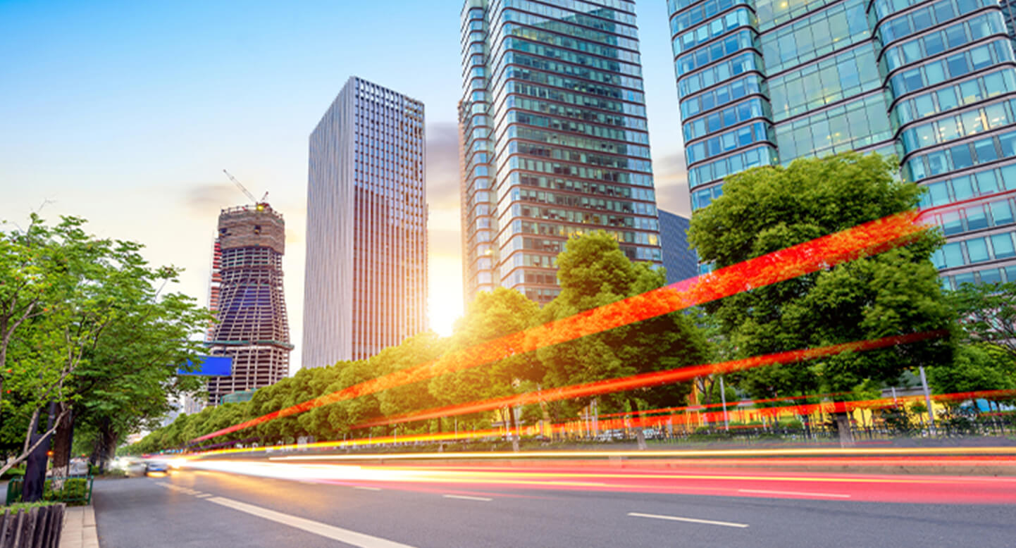 Streaks of light on a city street at sunset, with modern high-rise buildings and trees lining the road.