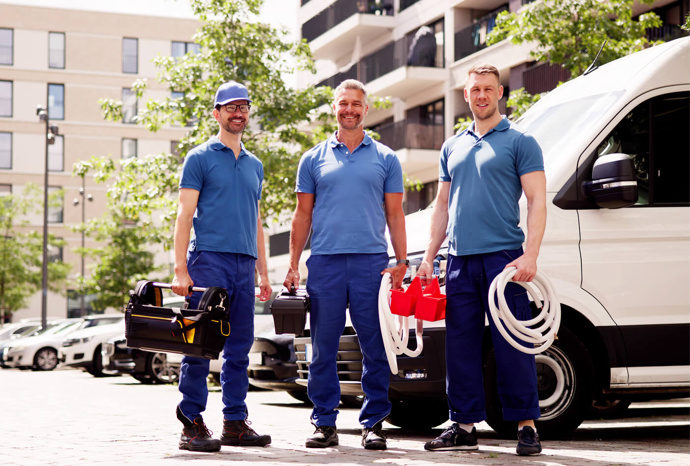 Three workers in blue uniforms hold tools and cables, standing by a white van in a sunny urban setting.