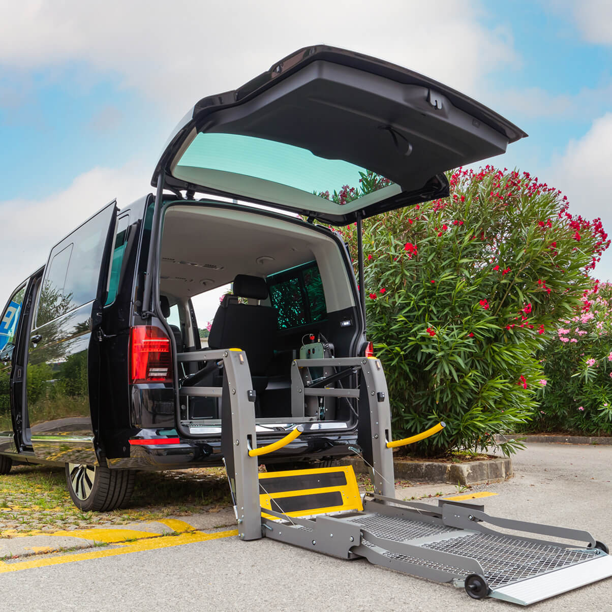 Black van with an extended wheelchair ramp, parked near green shrubs under a partly cloudy sky.