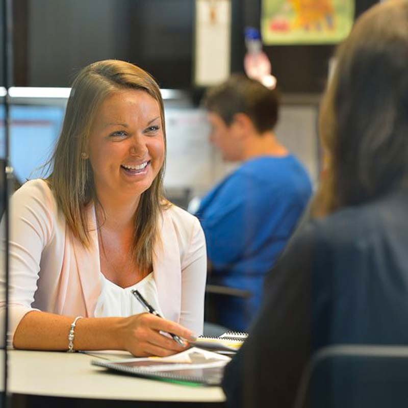 A woman smiling and holding a pen while talking to another person at a desk in an office setting.