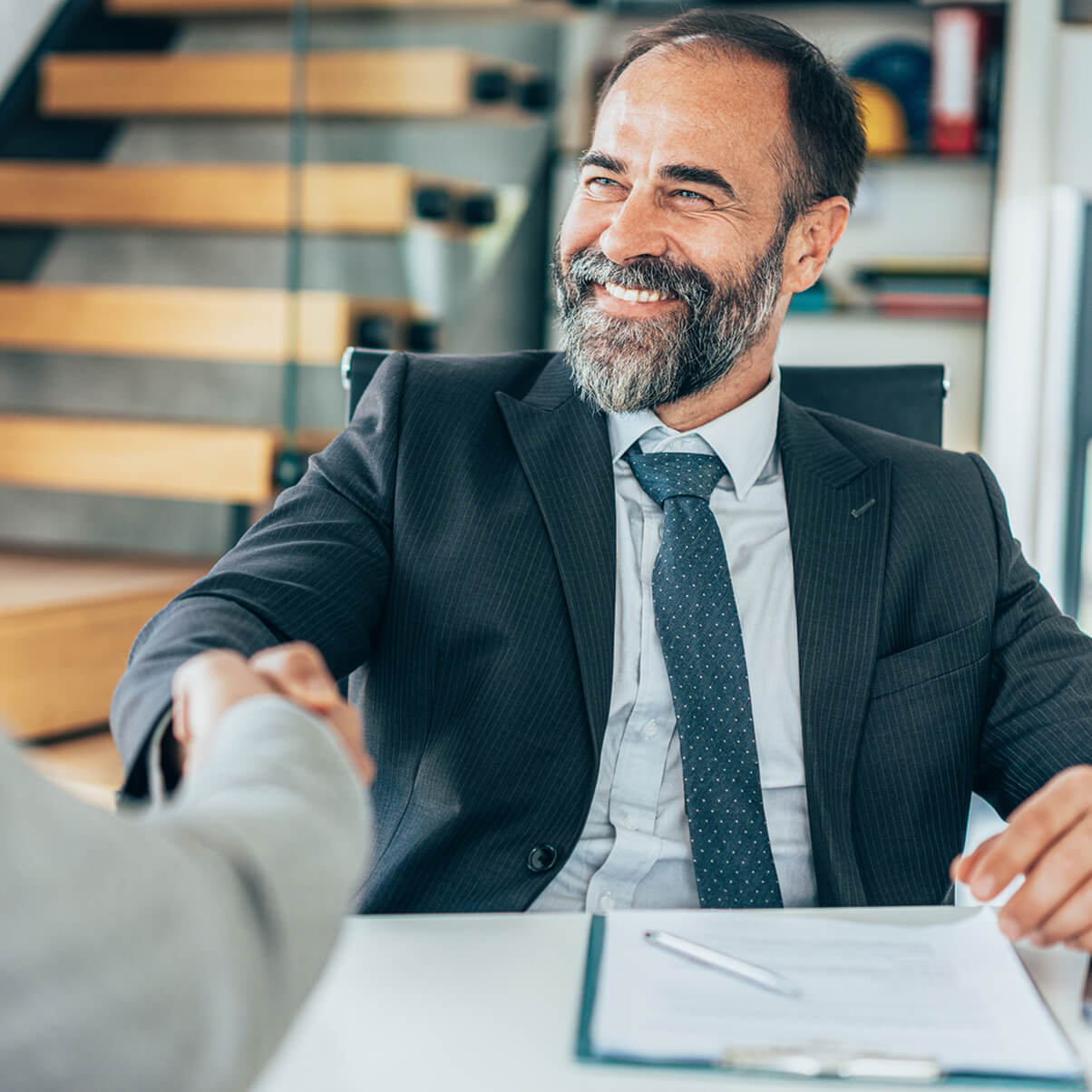 Man in a suit smiling, shaking hands with another person across a desk with a clipboard.