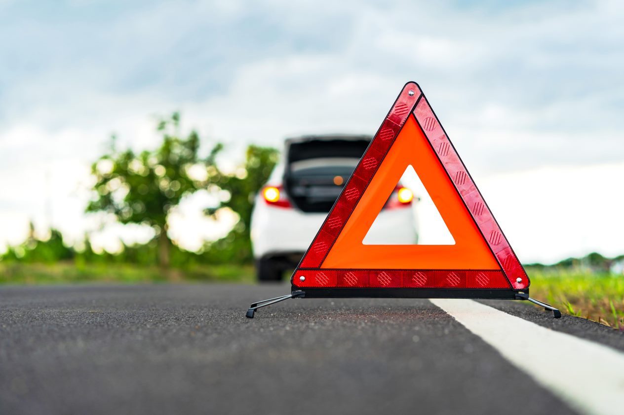 Red warning triangle on road with an open car trunk in background, green trees on a cloudy day.