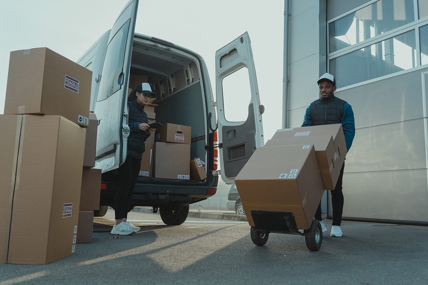 Two people load boxes into a van, with one using a hand truck outdoors near a building.