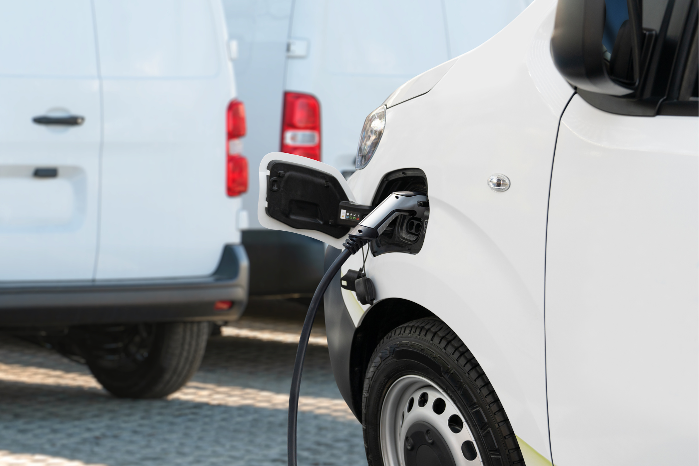 Electric van charging with a visible cable, parked on a cobblestone surface alongside other white vehicles.
