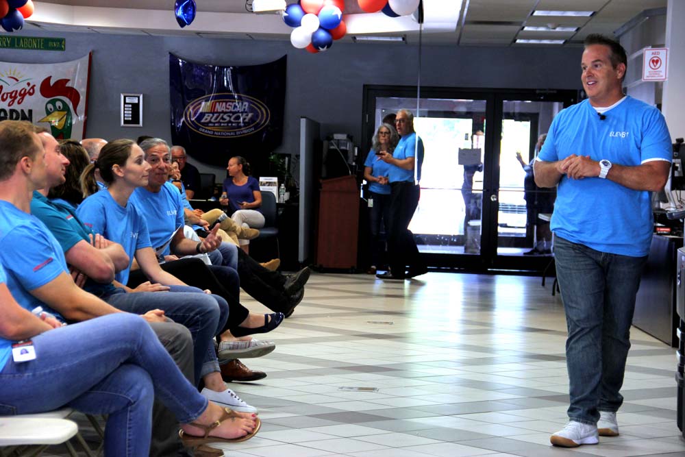 A man in a blue shirt speaks to an audience seated indoors, with balloons and banners decorating the room.