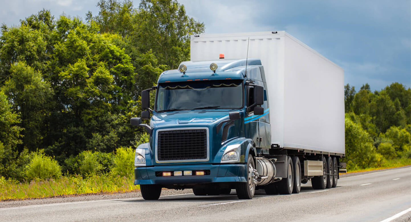 Blue semi-truck with a white trailer driving on a highway, surrounded by greenery and a cloudy sky.