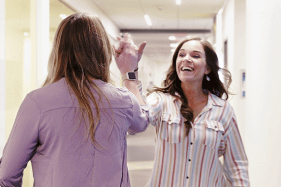 Two women high-fiving in a brightly lit hallway, both smiling and wearing casual shirts.